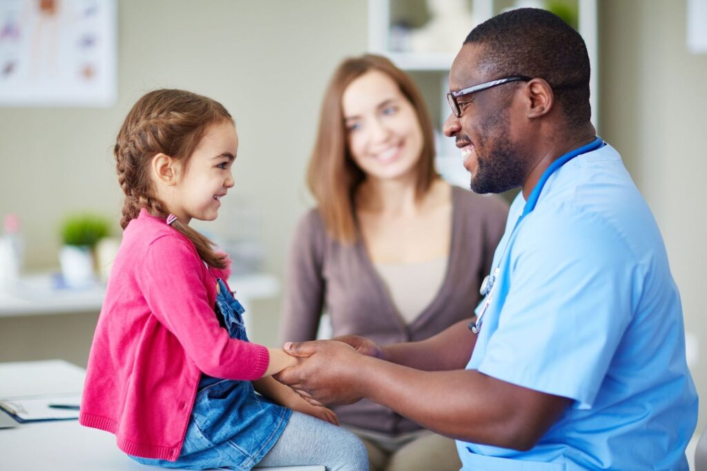 Smiling pediatric provider holding a young girl's hands while her mother looks on