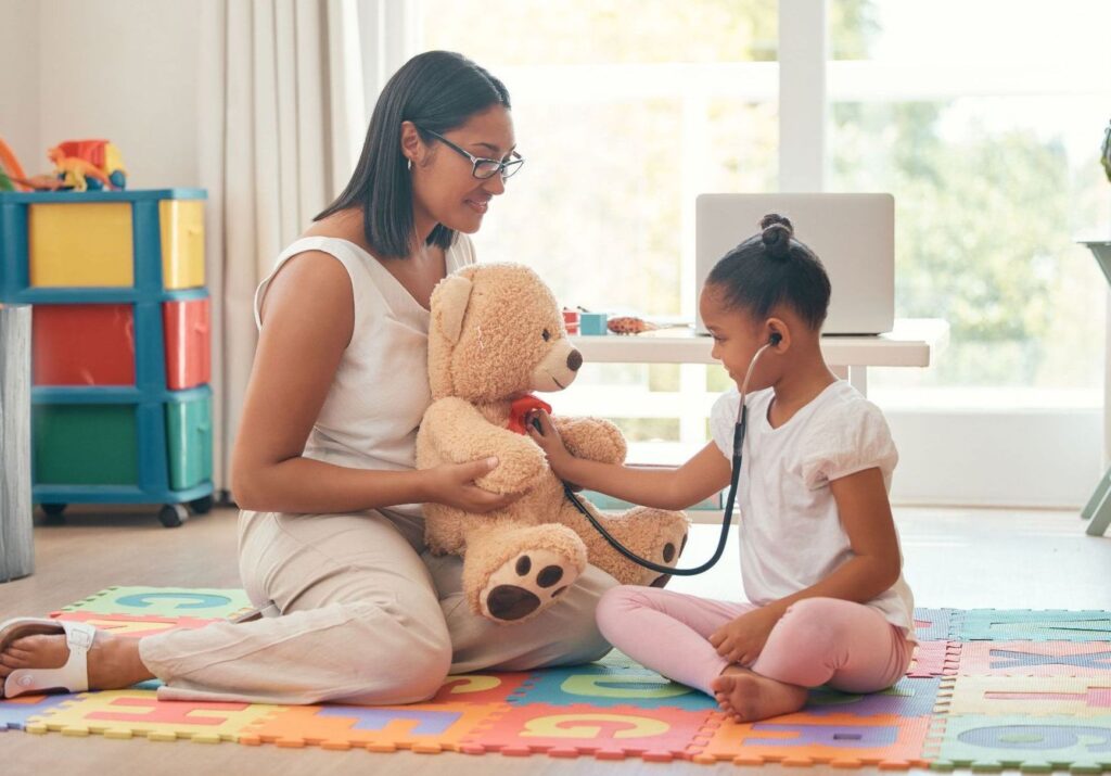 Young girl playing doctor, listening to a teddy bear's heartbeat with a stethoscope while a provider looks on