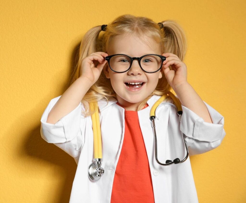 Smiling little girl in a doctor's coat and stethoscope adjusting her glasses against a yellow background