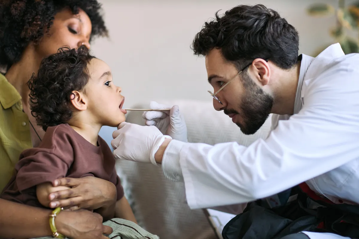 Pediatrician gently checking a toddler’s throat while the child sits in his mother’s lap